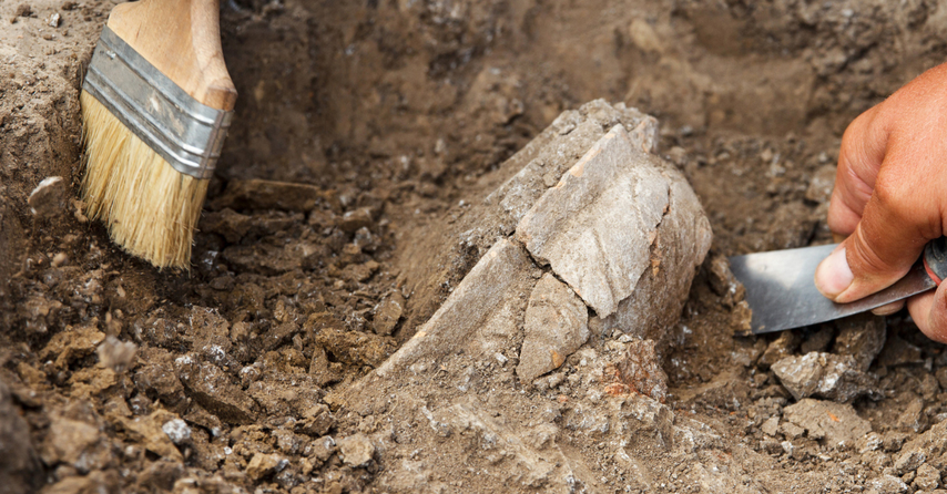 Close up of archeological dig, a brush in one hand and trowel in another digging in dirt.