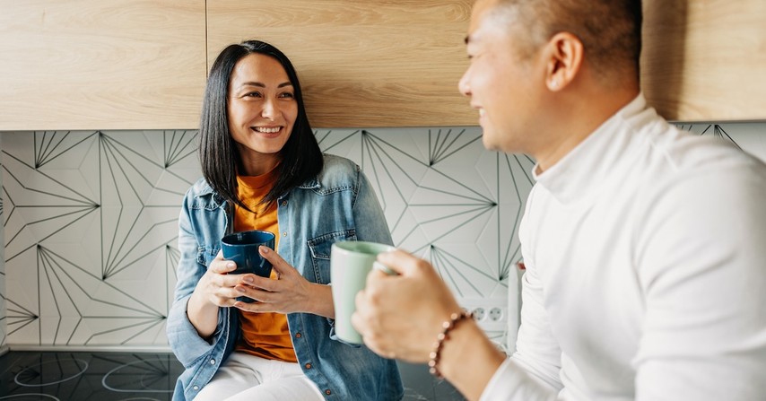 Happy asian couple in kitchen drinking coffee in mugs talking