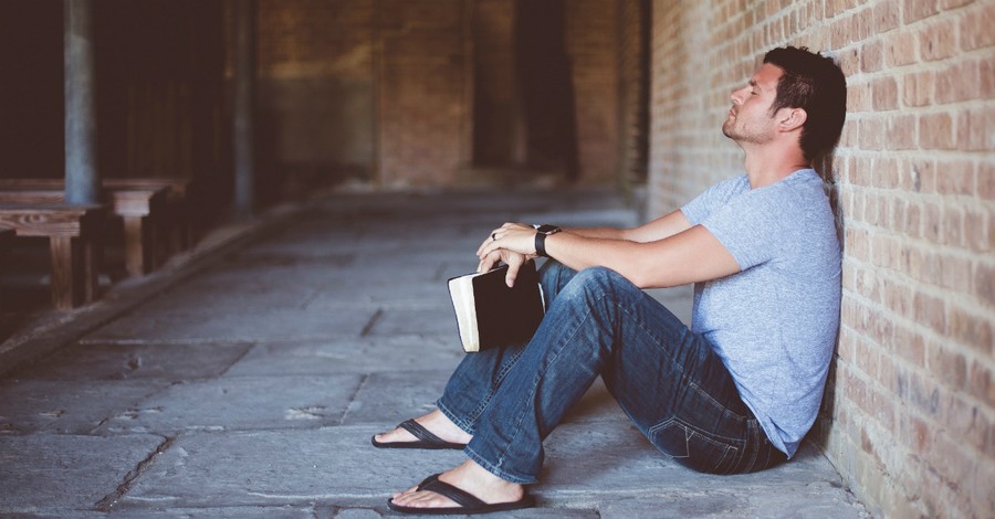 man sitting against wall with Bible praying